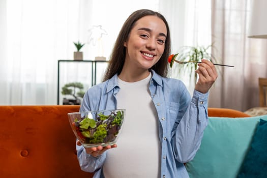Happy pregnant woman with great appetite eats vegetable salad sitting on sofa in cozy living room interior. Motherhood self-care childcare. Smiling Caucasian girl chooses the right healthy vegan food