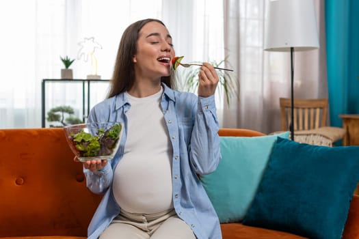 Happy pregnant woman with great appetite eating vegetable salad sitting on sofa in living room interior. Motherhood self-care childcare. Smiling Caucasian girl chooses the right healthy vegan food