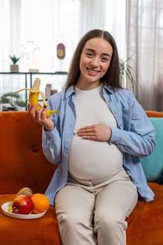 Happy pregnant woman with great appetite peels and eats banana fruit sits on sofa in living room with modern interior. Motherhood self-care childcare. Smiling girl chooses right healthy food. Vertical