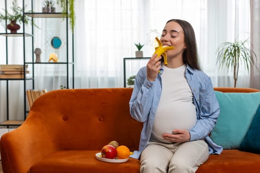 Happy pregnant woman with great appetite peels and eats banana fruit sits on couch in living room interior. Motherhood self-care childcare. Smiling Caucasian girl lady chooses right healthy food
