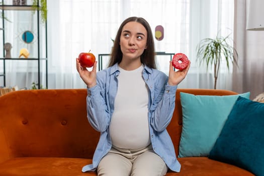 Choosing right nutrition. Confused young pregnant woman comparing sweet donut and ripe apple and shrugging shoulders in uncertainty. Future mother girl hesitates choosing between dessert and fruit.