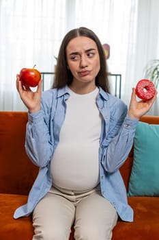 Choosing right nutrition. Confused pregnant woman comparing sweet donut and ripe apple and shrugging shoulders in uncertainty. Future mother lady hesitates choosing between dessert and fruit. Vertical