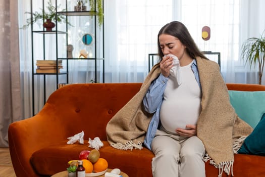 Pregnant unhealthy Caucasian woman girl wrapped in blanket blowing nose in tissue while sits on sofa couch in living room. Sick future mom feeling tired of nasal congestion and cold symptoms at home