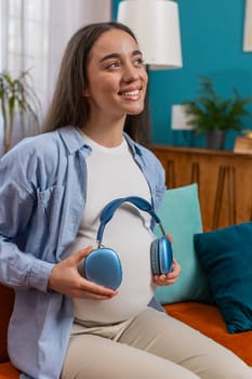 Happy pregnant woman sitting on sofa putting wireless headphones on her tummy with child baby while listening to favorite music at home room. Technology, future mother and domestic activity concept