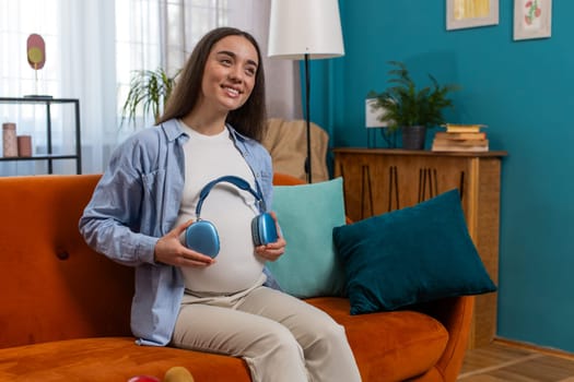 Happy pregnant woman sitting on sofa and putting wireless headphones on her tummy with child baby while listening to favorite music at home. Technology, future mother and domestic activity concept