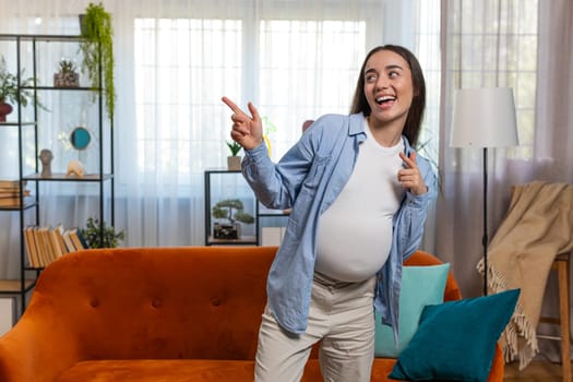 Joyful happy young funny Caucasian pregnant woman full of natural happiness and confidence enjoys playfully dancing and flashing friendly carefree smile. Future mother with big belly at home room.