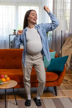Joyful young Caucasian pregnant woman full of natural happiness and confidence enjoys playfully dancing and flashing friendly carefree smile. Future mother with big belly at home room. Vertical