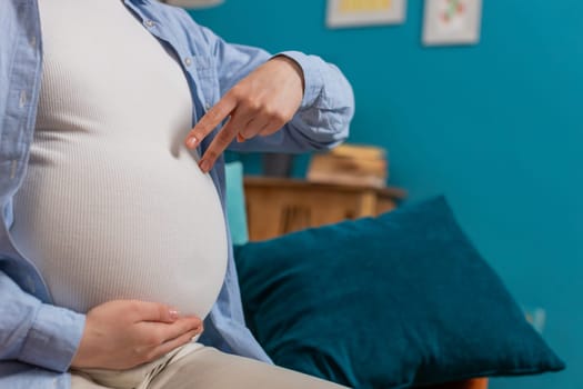 Close-up shot of unrecognizable young pregnant woman walks her fingers over her large belly sitting on sofa in living room at home. Future mother playing with unborn child in abdomen in apartment