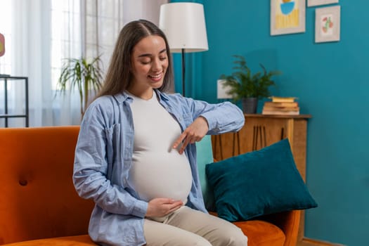 Happy beautiful young pregnant woman walks her fingers over her large belly sitting on sofa couch in living room at home. Smiling future mother playing with unborn child in abdomen in modern apartment