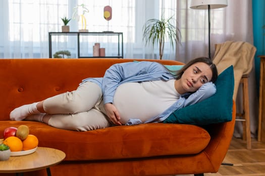 Upset young pregnant woman suffering from abdominal pain lying on sofa in living room at home. Illness future parent female relaxing in couch and feeling sick touching big belly in apartment.