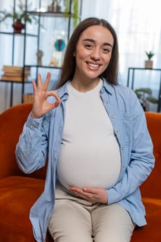Happy pregnant woman stroking big belly with love and showing ok sign sitting on sofa in living room at home. Pregnancy of young lady enjoy with future life, motherhood and pregnant concept.