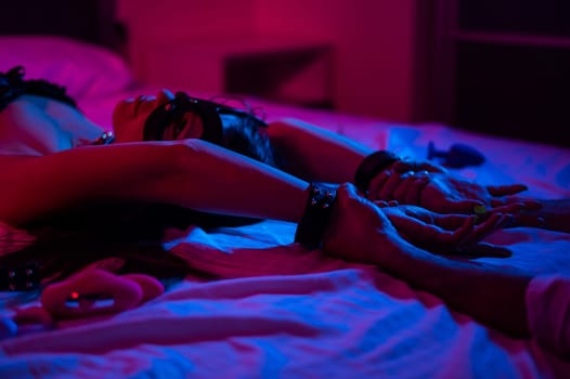 A woman in a leather mask and handcuffs lies on a bed in a blue-red light. Sex toys collection