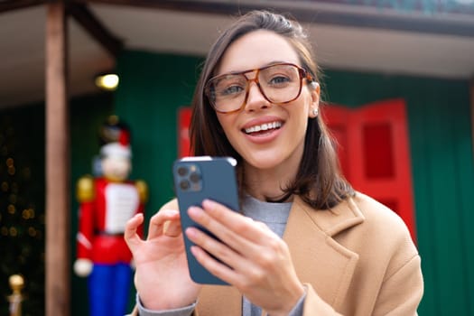 Woman in glasses using mobile phone at day in the city at Christmas holiday. Happy woman using smartphone dressed stylish trench coat looking at camera.
