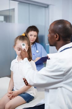 African American physician using thermometer on little girl to check fever in medical office. Doctor measures the temperature to find a diagnosis and gives treatment to the child in a cabinet.