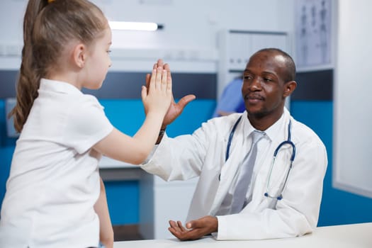 African american doctor giving high five to a young child during medical appointment in hospital office. Black man with a lab coat explaining illness symptoms discussing healthcare treatment.