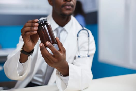 Close-up shot of a black man wearing a lab coat and holding a prescription medicine in the clinic office. Detailed image of African American doctor grasping a bottle of pills for recovery treatment.