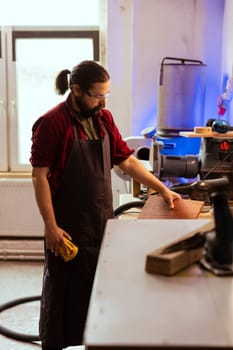 Artisan uses safety goggles to prevent workplace accidents while removing imperfections on wood. Man wearing protective equipment to avoid injury while using sandpaper to do sander plank