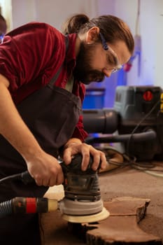 Cabinetmaker wearing protection glasses using angle grinder on wood to smooth surfaces by abrasion with sandpaper. Expert using orbital sander gear for furniture assembling job in carpentry shop