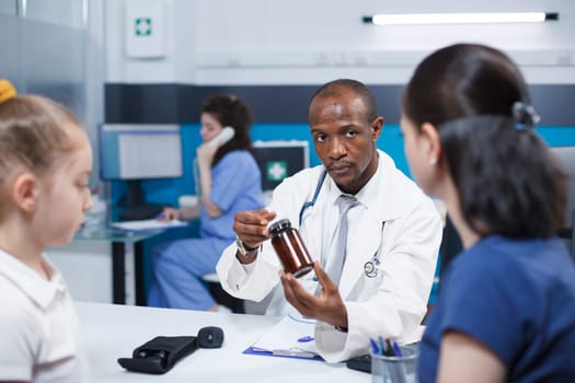 Mother and daughter consult with a healthcare professional about their concerns. African American physician explains treatment and diagnosis to Caucasian patients while holding medication bottle.
