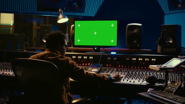 African american music producer working with greenscreen on pc, using mixing console to do mix and master in control room. Audio technician recording tracks with knobs and switchers. Camera B.