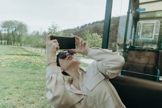 One beautiful young Caucasian brunette girl in sunglasses and stylish clothes takes pictures of nature on a mobile phone looking out of an open carriage of a tourist train on a spring sunny day in a public park, close-up side view.