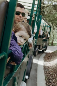 Portrait of one beautiful Caucasian little girl of two years old, who looks sweetly out of the open carriage of a tourist train and looks thoughtfully into the camera, driving through the reserve on a sunny spring day with her father, close-up side view.
