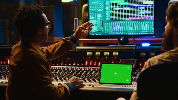 Professional engineer and musician mixing tracks next to isolated display on tablet, editing audio after recording session to produce a hit song. Artist collaborates with sound designer. Camera A.