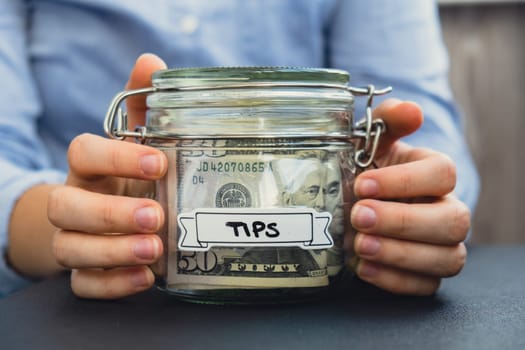 Female hands Saving Money In Glass Jar filled with Dollars banknotes. TIPS transcription in front of jar. Managing personal finances extra income for future insecurity background