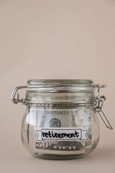 Saving Money In Glass Jar filled with Dollars banknotes. RETIREMENT transcription in front of jar. Managing personal finances extra income for future insecurity. Beige background
