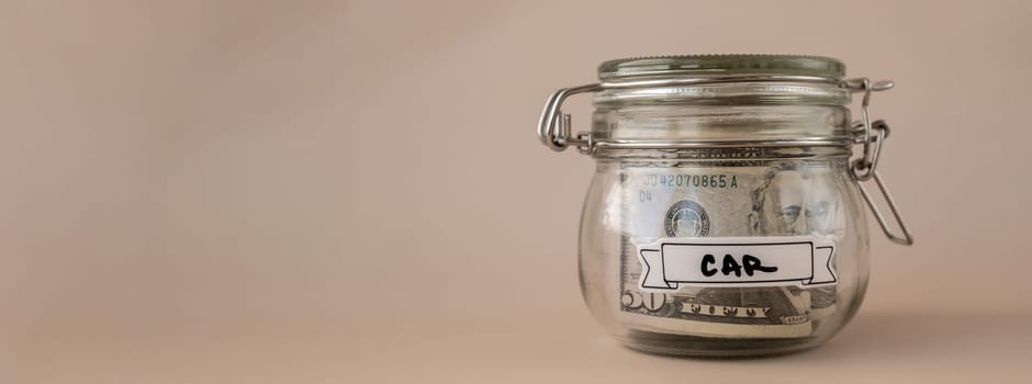 Saving Money In Glass Jar filled with Dollars banknotes. CAR transcription in front of jar. Managing personal finances extra income for future insecurity. Beige background