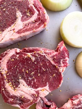 Three raw ribeye steaks with marbling, liberally seasoned with coarse spices, lie beside cut halves of onions on a kitchen tray, ready for cooking.