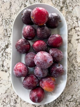 A plate of ripe, frosty plums sits prominently on a marble countertop in a sunlit kitchen with a crisp, white interior and neatly arranged appliances.