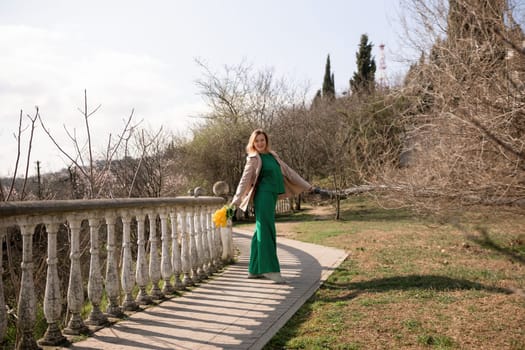 A woman in a green dress stands on a path next to a railing. She is holding a yellow bag and a yellow flower. The scene is peaceful and serene, with the woman posing for a photo