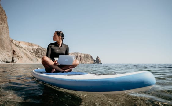 Digital nomad, woman in the hat, a business woman with a laptop sits on the rocks by the sea during sunset, makes a business transaction online from a distance. Freelance, remote work on vacation.