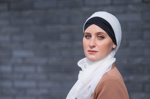 Portrait of a young blue-eyed woman in a hijab against a gray brick wall