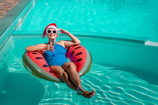 A happy woman in a blue bikini, a red and white Santa hat and sunglasses poses in the pool in an inflatable circle with a watermelon pattern, holding a glass of champagne in her hands. Christmas holidays concept