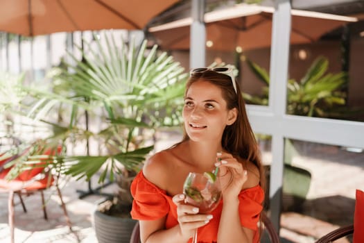 A woman in an orange dress sits at a table with a drink in a glass. The scene is set in a restaurant or cafe, with a potted plant nearby. The woman is enjoying her time, and the atmosphere is relaxed