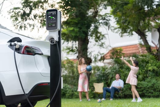 Focus electric vehicle recharge from home charging station on blur background of happy and playful family playing together. EV car using alternative and sustainable energy for better future.Synchronos