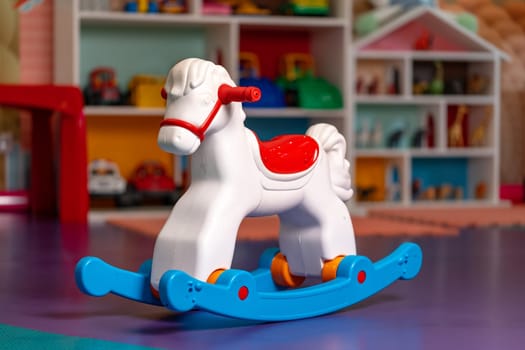 A plastic toy rocking horse rests on the floor of a playroom, surrounded by other childrens toys. The playroom is brightly lit, and the rocking horse is the focal point of the room.