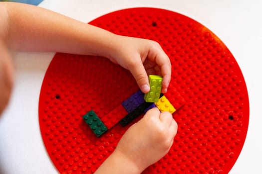 A young child is deeply focused while constructing a design with colorful plastic building blocks on a round red base. The childs hands are precisely placing a blue block to continue the pattern they have started, indicating an exercise in fine motor skills and creativity.