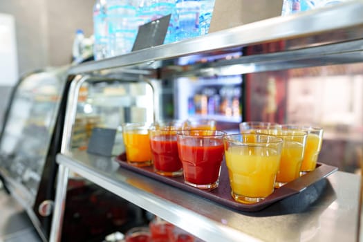 A selection of fruit juice cups, with orange and red hues, are neatly arranged on a metal serving cart, ready for patrons to enjoy in a bustling cafeteria setting. The focus on the front cups, with a blurred background, highlights the refreshment options available.