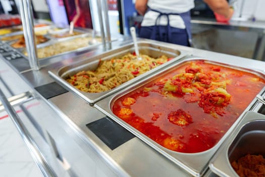 A bustling cafeteria serving line is filled with an array of dishes during a busy lunchtime. Trays of hot food are neatly arranged under the protective glass shield, with customers in the background selecting their meal choices. The variety suggests a diverse menu, catering to different tastes.