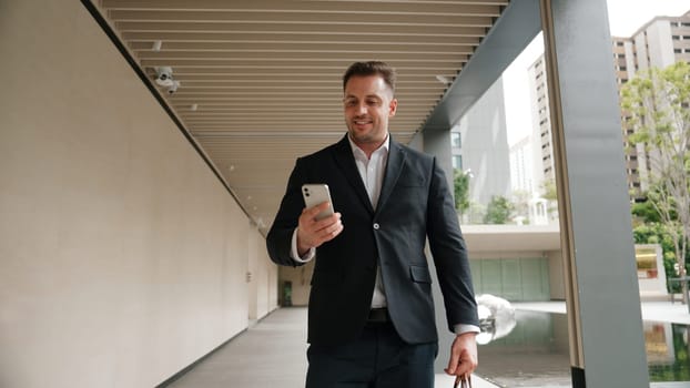 Happy project manager checking mobile phone while walking at corridor. Professional business man celebrate successful project or increasing sales while wear suit and hold suitcase for meeting. Urbane.