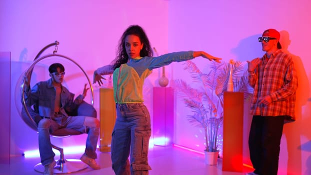 Attractive happy dancer looking at camera while smart woman moving at rhyme with neon light. Professional hispanic performer break dancing while wearing colorful cloth with diverse friend. Regalement.