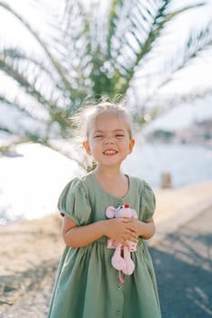 Little smiling girl with a toy pink mouse in her hand stands on the seashore near a green palm tree. High quality photo