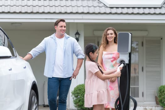 Happy little young girl learn about eco-friendly and energy sustainability as she help her family recharge electric vehicle from home EV charging station. EV car and modern family concept. Synchronos