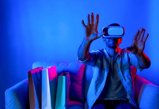 Caucasian man with vr glass posing pointing finger while sitting at sofa with shopping bags. Person selecting and doing shopping online by using VR technology and virtual goggle at home. Deviation.