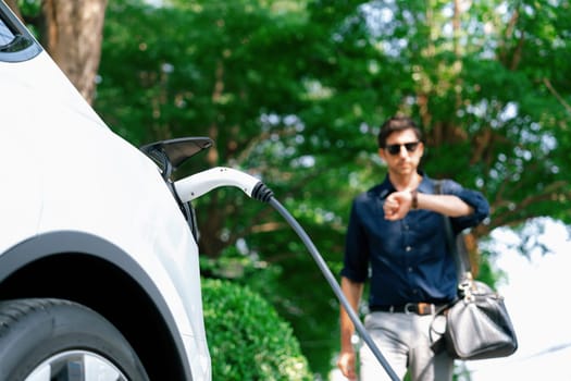 Young man recharge electric car's battery from charging station in outdoor green city park in springtime. Rechargeable EV car for sustainable environmental friendly urban travel lifestyle. Expedient