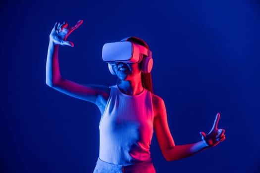 Female stand surrounded by neon light wear VR headset connecting metaverse, futuristic cyberspace community technology, spread both hand index and thumb finger interact virtual object. Hallucination.