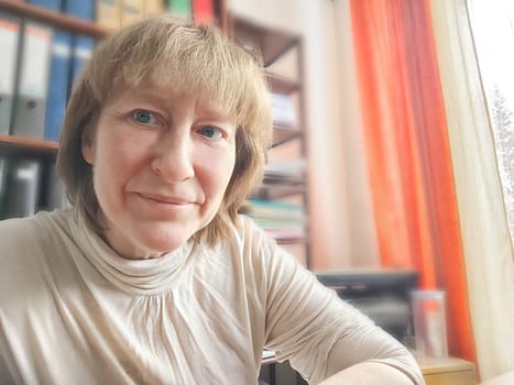 Funny blonde girl or a middle-aged woman poses for the camera with a smile and closet with folders on background. A relaxed Portrait of an Accountant, manager, blogger, freelancer in the workplace